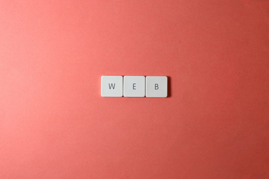 A close-up photograph features three white square tiles with black letters spelling 'WEB' arranged in a horizontal line on a smooth, matte pink background. The tiles have a slightly raised edge and ro